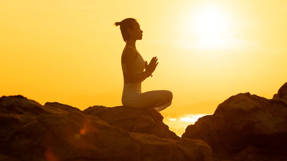 A woman meditates in a yoga pose on rocks during a warm, golden sunset.